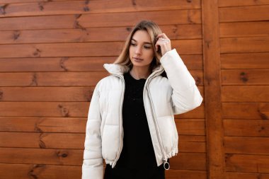 Şehrin dışında, ahşap bir binanın yanında siyah kazaklı, şık beyaz ceketli hoş bir genç kadın modeli. Güzel, şık bir kız, modaya uygun kıyafetlerle sokakta yürüyor. Bahar stili.