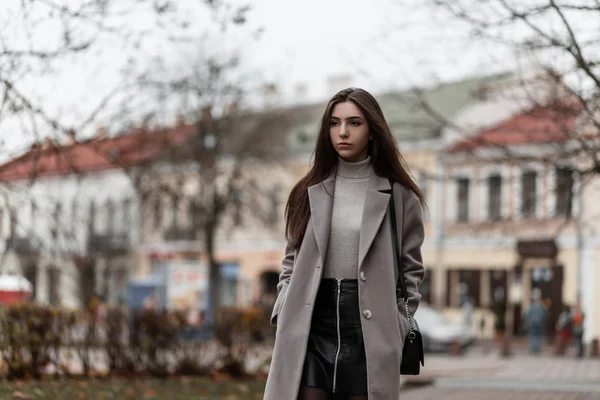 Jeune femme élégante avec les cheveux longs dans un manteau d'élégance dans une jupe noire avec un sac à main en cuir à la mode se tient et profite d'un week-end dans la rue dans la ville. Mignon fille modèle en plein air en automne jour — Photo