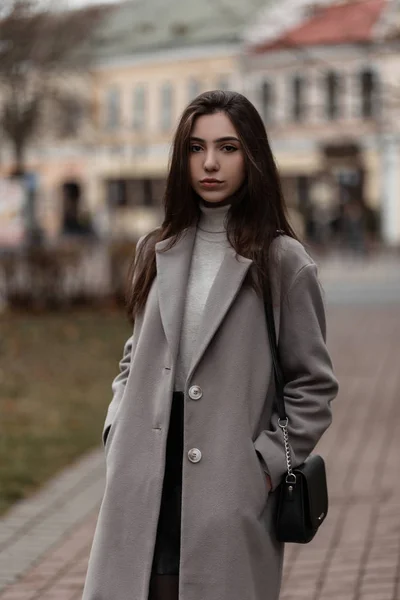 Μοντέρνο μοντέλο μιας νεαρής γυναίκας με καστανά μαλλιά σε ένα κομψό παλτό σε ζεστό vintage γκολφ με τσάντα που θέτουν στην πόλη σε μια ημέρα του φθινοπώρου. Μοντέρνο κορίτσι στέκεται στο δρόμο. Άνοιξη-φθινόπωρο στυλ. — Φωτογραφία Αρχείου