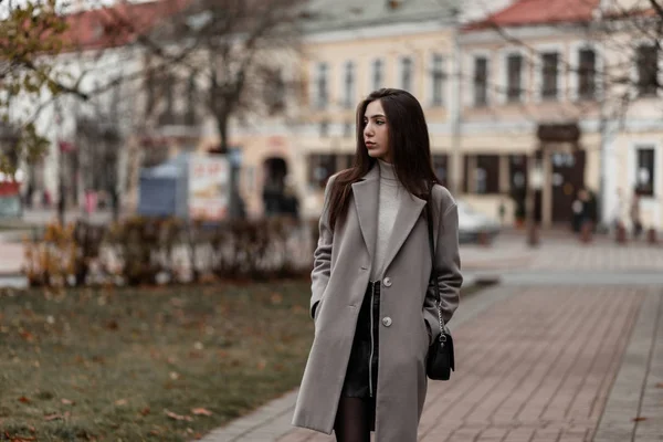 Mulher jovem europeia em um casaco cinza na moda com uma bolsa de couro preto elegante fica em uma rua na cidade em um dia de outono. Menina morena bonito goza de uma caminhada ao ar livre . — Fotografia de Stock