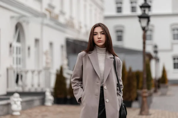 Moderna giovane donna carina in un elegante cappotto con una borsetta di pelle in una gonna nera cammina lungo la strada vicino a vintage edificio bianco. Ragazza urbana europea in abiti alla moda in città in una giornata autunnale . — Foto Stock