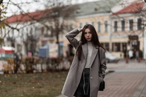 Atraktivní hezká mladá žena v elegantním kabátě v černé sukni s módní koženou taškou kráčí ulicí. Elegantní městská dívka ve stylovém oblečení ve městě venku. Podzimní ležérní svrchní oděv. — Stock fotografie