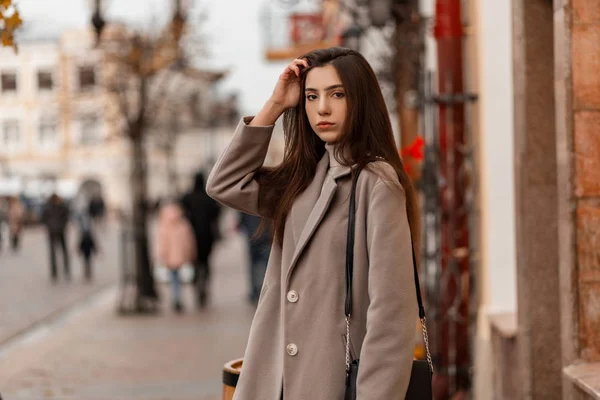 Joven modelo de moda hermosa mujer con pelo largo marrón en un abrigo de otoño gris de moda con un bolso de cuero negro posando cerca de un edificio vintage en la ciudad.Muchacha atractiva descansando en la calle — Foto de Stock