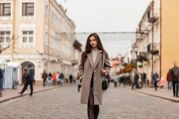 Mulher europeia jovem em um casaco na moda em uma saia de couro no golfe quente com uma bolsa preta elegante caminha pelas ruas da cidade perto de edifícios em um dia de outono. Modelo menina atraente ao ar livre . — Fotografia de Stock