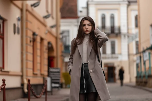 Leuke jonge vrouw met lang bruin haar in een modieuze jas in een leren vintage rok met een zwarte trendy handtas die poseert in de stad. Mooi meisje loopt in de buurt van moderne gebouwen.Jeugd dameskleding — Stockfoto