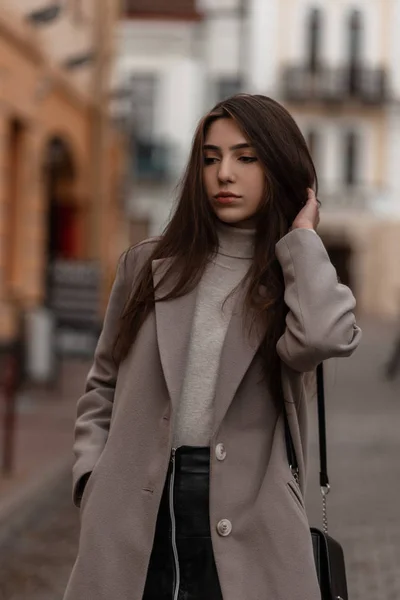 Retrato de uma jovem menina bonita com cabelos longos chiques em um elegante casaco de outono com uma bolsa preta na moda na rua perto de edifícios vintage. Modelo de moda menina elegante atraente ao ar livre . — Fotografia de Stock