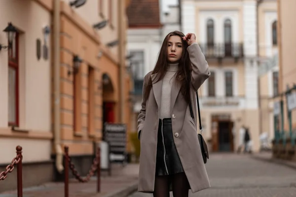 Mujer joven bastante atractiva con el pelo castaño en un abrigo de lujo en una falda de cuero con una bolsa de moda disfruta de un paseo por la calle en un día de otoño. Hermosa chica en la ropa elegante al aire libre . — Foto de Stock