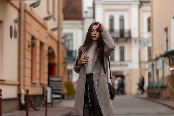 Bonito modelo de uma jovem mulher em um casaco elegante em uma saia de couro elegante no golfe vintage com uma bolsa preta na moda posa perto de um edifício na cidade. Menina bonita anda ao redor da rua . — Fotografia de Stock