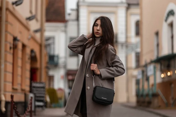 Mulher morena jovem bonita em um casaco longo na moda com uma bolsa preta de couro elegante posa na cidade perto de um edifício vintage. Urbano bonito menina ao ar livre. Roupa exterior feminina sazonal na moda . — Fotografia de Stock