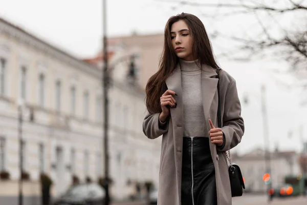 Mulher jovem urbana com cabelos longos em um casaco elegante em uma saia preta com uma bolsa de couro elegante fica e goza de um fim de semana na rua na cidade.Modelo menina bonita ao ar livre no dia de outono — Fotografia de Stock