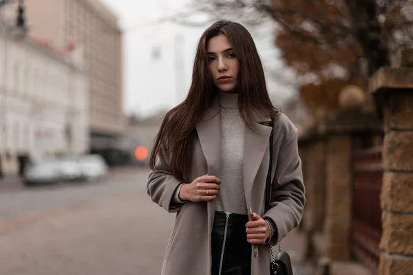 Mulher muito jovem com cabelos longos em um casaco elegante em uma saia preta com uma bolsa de couro elegante fica e goza de um fim de semana na rua da cidade. Bonito modelo menina ao ar livre no dia de outono . — Fotografia de Stock