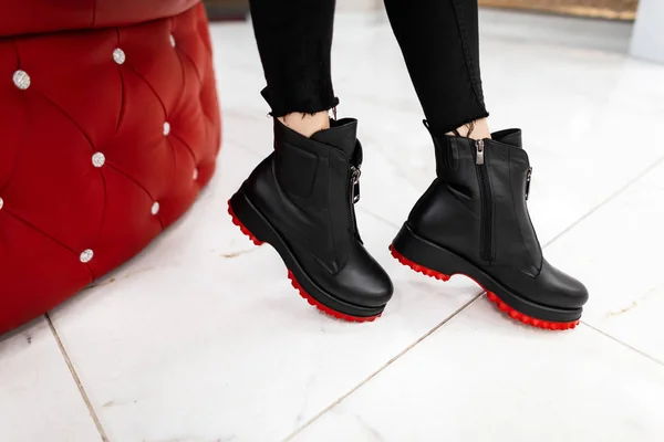 Jeune femme moderne en jean élégant noir en bottes en cuir à la mode avec une semelle rouge est debout sur une tuile dans une pièce près d'un fauteuil rouge doux. Collection tendance de chaussures d'automne pour femmes. Gros plan . — Photo