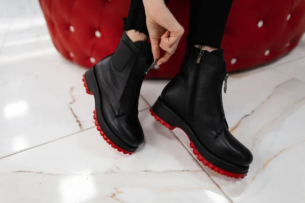 Módní žena sedí v obchodě a měří podzimní boty.Detailní záběr ženských nohou ve stylových džínách v módních kožených černých botách s červenými podrážkami. Nová sezónní kolekce dámské obuvi. — Stock fotografie