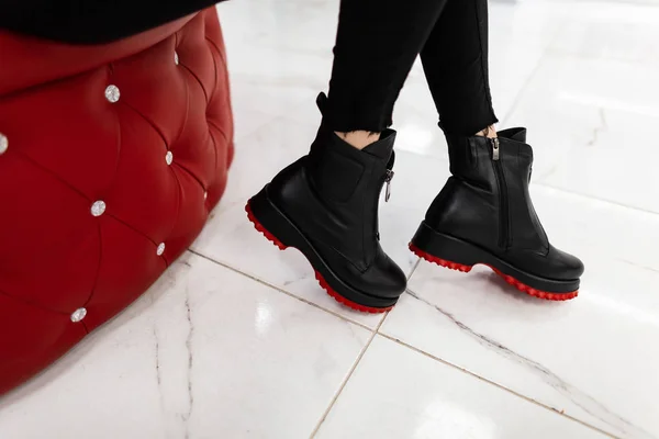 Detailní záběr ženských nohou v módních kožených černých botách s červenými podrážkami ve starých džínách uvnitř. Stylová dívka v módních botách sedí na měkkém křesle v obchodě. Nakupování — Stock fotografie