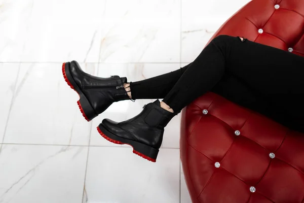 Модные женские сезонные туфли. Новая осенняя стильная коллекционная обувь. Вид сверху на женские ноги в черных джинсах в кожаных трикотажных черных сапогах лежит на красном роскошном диване — стоковое фото