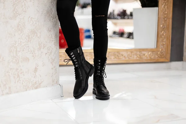 Módní dámské sezónní boty. Nová kolekce. Mladá žena ve stylových černých kožených botách ve starých džínách stojí ve světlé místnosti. Nakupovat. Detailní záběr. — Stock fotografie