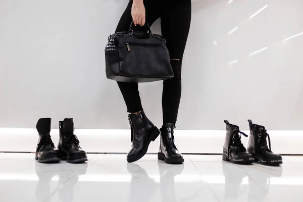 Модная новая коллекция сезонной женской обуви и сумок. Крупный план женских ног в стильных черных джинсах в сапогах с кожаной винтажной сумкой в светлой комнате. Девушка выбирает модную обувь . — стоковое фото