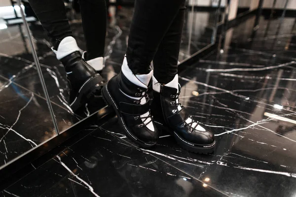 Closeup of female legs in fashionable warm leather black-white boots on  in vintage jeans indoors. Stylish girl in trendy shoes stands on the tile near the mirror in the store. Autumn-winter style. — Stock Photo, Image