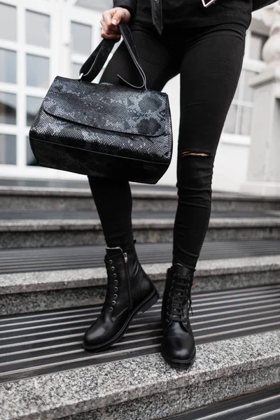 Σύγχρονη νεαρή γυναίκα με μια μοντέρνα μαύρη δερμάτινη τσάντα σε τζιν το φθινόπωρο κομψό δερμάτινες μαύρες μπότες στέκεται στο δρόμο. Απλή μοντέρνα νεανική εμφάνιση. Κοντινό πλάνο γυναικείων ποδιών. — Φωτογραφία Αρχείου
