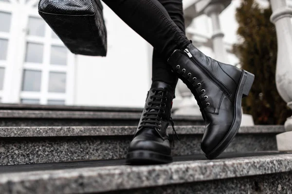 Jonge trendy vrouw in stijlvolle lederen zwarte laarzen in jeans met een tas staat op de stenen treden in de stad. Close-up van vrouwelijke benen in mode schoenen. Nieuwe collectie schoenen en accessoires. — Stockfoto