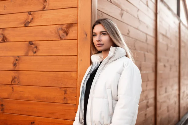 Hermoso modelo urbano de una mujer joven con el pelo largo rubio en ropa negra con estilo en una chaqueta blanca de moda posa cerca de una pared de madera vintage en la ciudad. Linda chica con labios sexy en la calle . — Foto de Stock