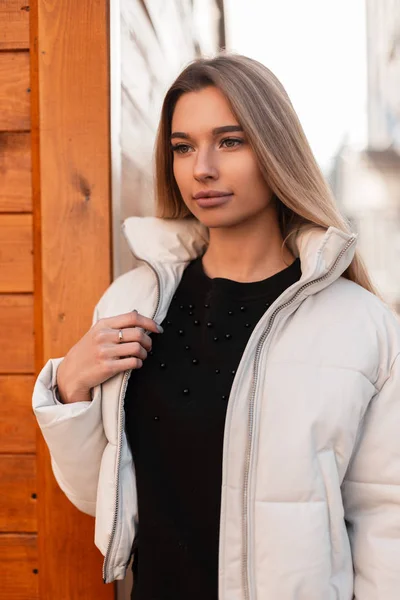 Ritratto di una bella giovane donna con i capelli biondi in una giacca bianca alla moda in un maglione nero vintage per strada vicino a un edificio di legno. Attraente ragazza moderna con trucco naturale in città . — Foto Stock