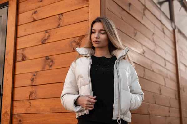 Jolie jeune femme européenne blonde dans une veste de printemps blanche vintage dans un élégant pull noir posant près d'un bâtiment en bois brun dans la rue. À la mode jolie fille urbaine mannequin de mode en plein air . — Photo