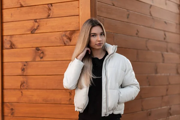 一个穿着时尚的白色夹克，穿着黑色毛衣的年轻貌美的女人在一座木头建筑附近的城市的模型。 穿着时髦衣服的漂亮女孩在街上走着。 春风. — 图库照片