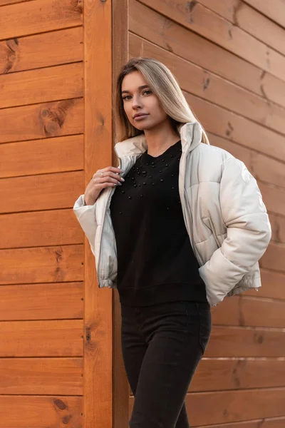 Νέοι ελκυστική ξανθιά γυναίκα σε ένα μαύρο πουλόβερ σε ένα μοντέρνο λευκό σακάκι στέκεται στο δρόμο κοντά σε ένα καφέ ξύλινο κτίριο. Χαριτωμένο κορίτσι με μοντέρνα ρούχα απολαμβάνει μια βόλτα στην πόλη. Στυλ. — Φωτογραφία Αρχείου