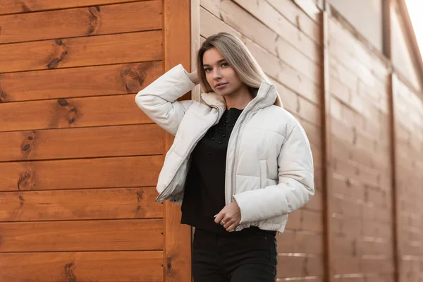 Jolie jeune femme blonde glamour avec des lèvres sexy dans un pull élégant noir dans une veste d'hiver blanche à la mode posant à l'extérieur près d'un bâtiment vintage en bois. Une fille debout en ville. Mode jeunesse . — Photo