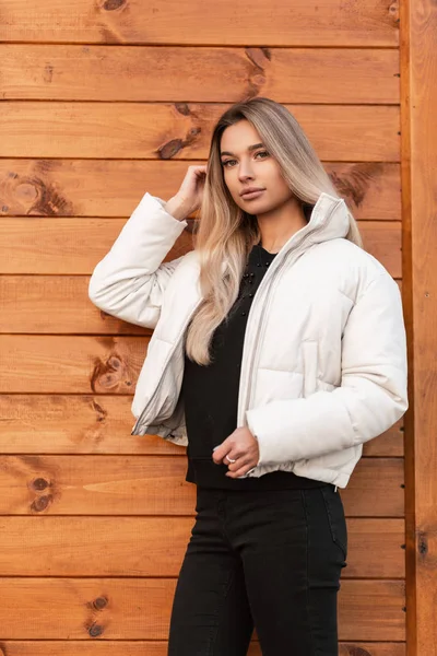 Hermoso modelo moderno de una mujer joven con el pelo largo rubio en ropa negra con estilo en una chaqueta blanca de moda posa cerca de una pared de madera vintage en la ciudad. chica urbana con labios sexy en la calle — Foto de Stock