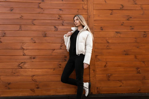 Jovem mulher elegante loira em botas de couro branco na moda em uma jaqueta elegante em jeans pretos relaxa na rua perto de um edifício vintage de madeira. Menina da moda atraente está descansando ao ar livre . — Fotografia de Stock