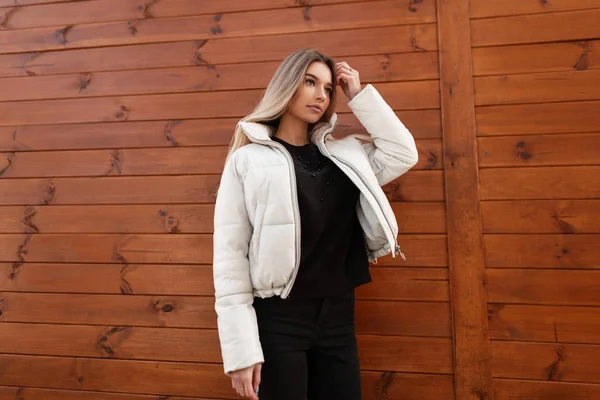 Mulher loira atraente muito jovem em uma camisola preta em uma jaqueta branca na moda fica na rua perto de um edifício de madeira marrom. Menina urbana em roupas da moda está desfrutando de um passeio na cidade . — Fotografia de Stock