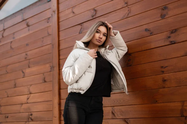 Bellissimo modello glamour di una giovane donna con i capelli lunghi biondi in eleganti vestiti neri in una giacca bianca alla moda posa vicino a un muro di legno vintage in città. bionda ragazza urbana riposa in strada . — Foto Stock