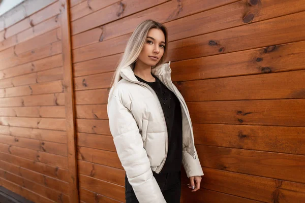 European glamorous young woman blonde in a fashionable white jacket in black jeans in a sweater stands outdoors near a wooden vintage wall. Attractive modern girl fashion model walks on the city. — Stock Photo, Image