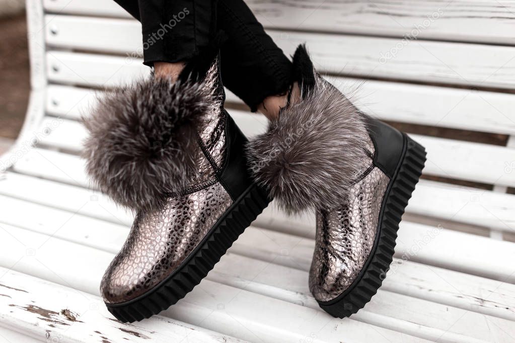 Fashionable silver shoes with gray fur on female legs. Stylish young woman in a glamorous shiny winter shoes sits on a wooden bench in the city. Close-up of trendy seasonal warm women's footwear.