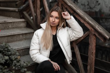 Beyaz, modaya uygun, siyah kot pantolonlu, sokakta ahşap korkuluklu eski bir merdivende poz veren şık, genç bir bayan. Moda kıyafetleri içinde güzel bir kız model.