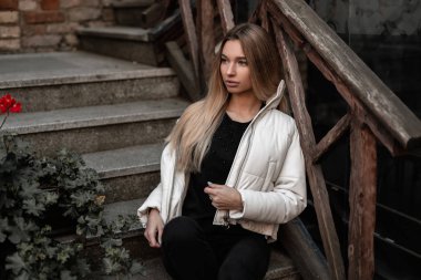 Modaya uygun siyah-beyaz dış giyimli güzel bir kadın model, tahta korkuluklu eski bir merdivende dinleniyor. Şık giyimli çekici kız şehir dışında dinleniyor. Sonbahar