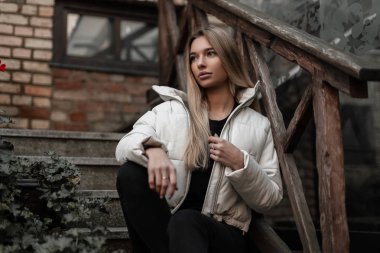 Beyaz ceketli, siyah kotlu bir süveter giyen, klasik ahşap korkuluklu taş bir merdivende oturan Avrupalı genç bir kadın. Moda kıyafetleri içinde sevimli sarışın kız..
