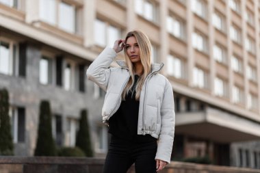 Gençlik modasına uygun giyinen modern sarışın genç kadın saçları düzleştiriyor. Beyaz modaya uygun sezonluk ceketli şık bir manken şehrin caddesinde klasik bir binanın yanında poz veriyor..
