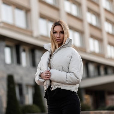 Modern bir binanın arka planında şık beyaz ceketli güzel, modaya uygun genç bir kadın duruyor. Havalı sonbahar-bahar kıyafetleriyle dışarıda poz veren çekici kız mankeni..
