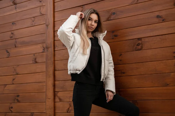 Mulher jovem europeia bonito em uma jaqueta branca na moda em jeans preto elegante em um suéter de malha posa perto de uma parede vintage de madeira. Menina loira moderna atraente goza de um passeio na cidade . — Fotografia de Stock