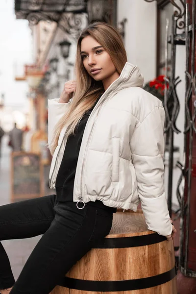 Молодая привлекательная женщина с длинными светлыми волосами в модной белой куртке в черных джинсах в трикотажном свитере сидит на деревянной винтажной бочке на открытом воздухе. Красивая девушка-модель отдыхает на улице. Мода . — стоковое фото