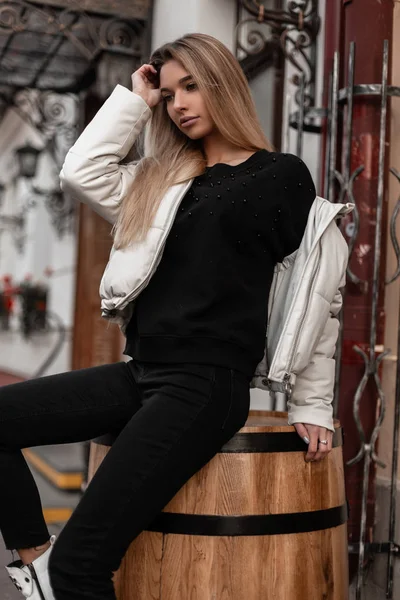 Mulher loira jovem europeia em uma jaqueta branca na moda em jeans elegantes pretos em um suéter vintage de malha está descansando sentado em um barril de madeira na cidade. Menina na moda ao ar livre em um dia de outono . — Fotografia de Stock