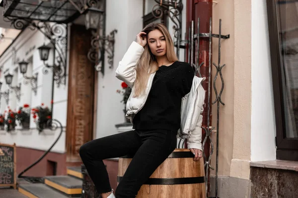 Χαριτωμένο μοντέλο μιας νεαρής όμορφης γυναίκας με μοντέρνα εξωτερικά ενδύματα κάθεται σε ένα παλιό ξύλινο βαρέλι κοντά σε ένα κτίριο στο δρόμο. Ευρωπαϊκό κομψό κορίτσι με μοντέρνα ρούχα ξεκουράζεται σε εξωτερικούς χώρους. Άνοιξη-φθινόπωρο — Φωτογραφία Αρχείου