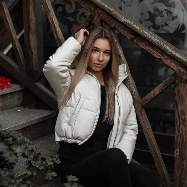 Mulher loira jovem bonita em uma jaqueta branca na moda em jeans elegantes pretos está descansando em uma escada vintage de pedra na rua. Menina europeia na moda está sentada nos degraus da cidade . — Fotografia de Stock