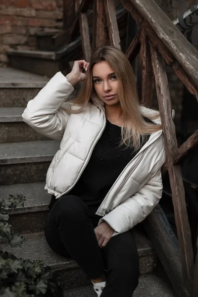 Mujer joven con estilo urbano en una chaqueta blanca de moda en vaqueros negros vintage posando sentado en una vieja escalera de piedra con barandilla de madera en la ciudad. Linda chica modelo en ropa de moda en la calle — Foto de Stock