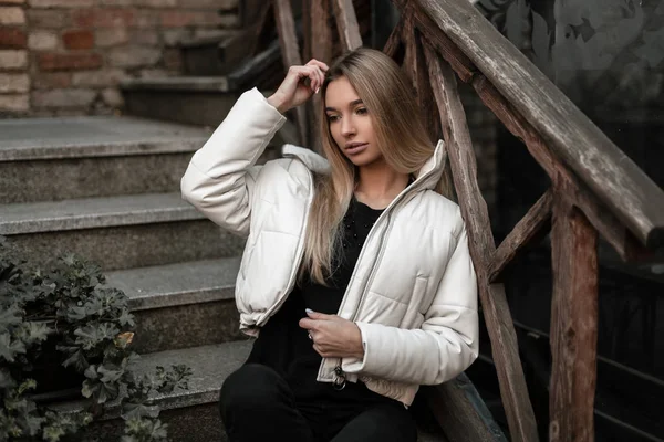 Jovem na moda em roupas da moda com longos cabelos loiros está descansando na cidade em uma escada de pedra com corrimão de madeira vintage. Modelo menina bonita em um outerwear elegante ao ar livre. Outono . — Fotografia de Stock