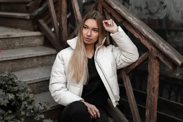 Cute stylish young woman in a white fashionable jacket in vintage black jeans posing sitting on an old stone staircase with wooden railing on the street.Beautiful girl model in trendy clothes outdoors — Stock Photo, Image