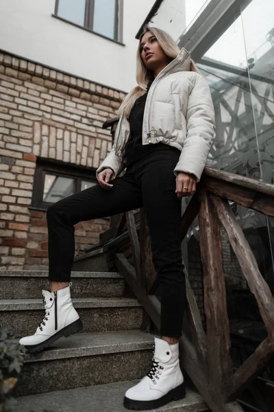 Λεπτή όμορφη νεαρή γυναίκα με μαύρο τζιν σε ένα μοντέρνο λευκό σακάκι σε δερμάτινες λευκές μπότες στέκεται σε μια πέτρινη σκάλα στην πόλη. Όμορφη σύγχρονη κοπέλα αναπαύεται κοντά σε ένα ξύλινο κιγκλίδωμα σε εξωτερικούς χώρους. — Φωτογραφία Αρχείου